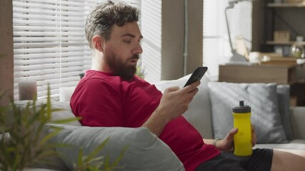 Tired guy drinking water, taking break in workout at home. Sport and hydration. Siting on sofa - Powered by Adobe