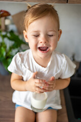 Happy baby girl sitting at the table in the kitchen and drinking milk