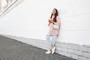 Beautiful young pregnant woman wearing pink sweater, jeans and a bag standing near white wall on a...