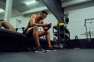 Fototapeta na wymiar Young African American male looking at his phone to reply to an online message. Happy, mixed race male smiling at his phone while sitting down in the gym. High quality photo 