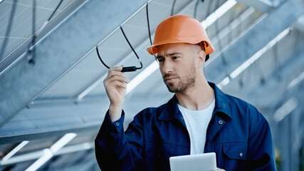 engineer in hardhat checking wire of solar panels while holding digital tablet