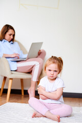 Fototapeta na wymiar Upset little girl sitting lonesome while mother using laptop. Mother don't have time for daughter