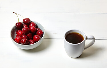 White cup with coffee and sweet cherries in a ceramic bowl on a white wooden background, closeup. Tasty breakfast, morning routine concept