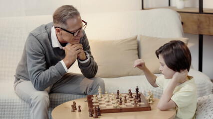 grandfather in glasses looking at smart grandson while playing chess in living room