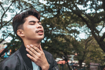A young man experiences a sore or scratchy throat while at the park. Outdoor park scene - obrazy, fototapety, plakaty