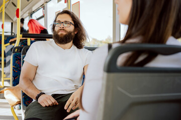 Man with long hair and thick beard wearing glasses takes public transport bus to work to meeting with friends looks out window observes bus stops afternoon in large agglomeration traffic jam rush hour - obrazy, fototapety, plakaty