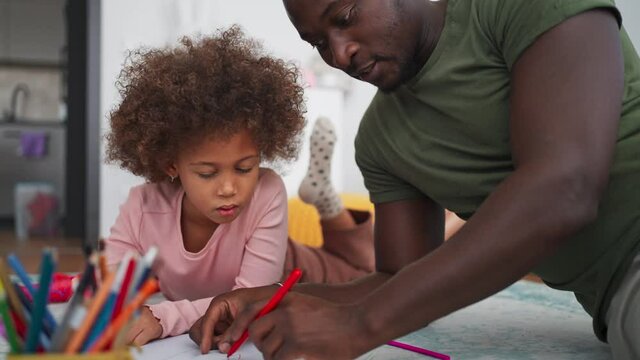 Little multiracial girl lying on floor and doing homework with his african american father at home.