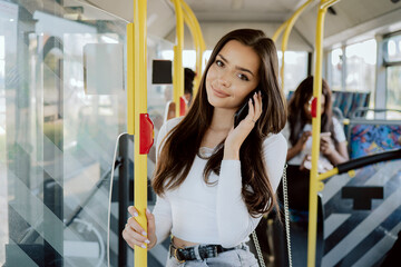 A bus trip in the city, returning from work, a student's way to university, a beautiful girl is riding public transport, holding on to the railing at the door, talking on the phone, smiling at camera