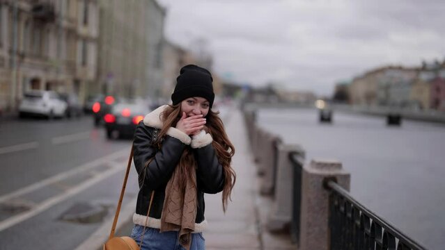 happy charming woman is warming hands by breathing in city street, walking alone in quay in winter day