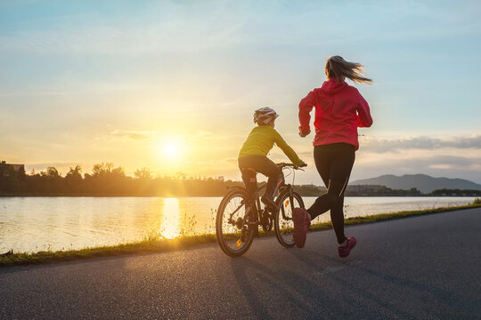 Happy mother and son go in sports outdoors. Boy rides bike in helmets, mom runs on sunny day. Silhouette family at sunset. Fresh air. Health care, authenticity, sense of balance and calmness
