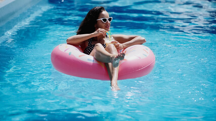 happy woman in stylish sunglasses holding glass with cocktail and swimming on inflatable ring in pool