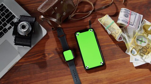 Mobile phone and smartwatch with chroma key top view, money cash, compass, photo camera and laptop computer on table. Traveling and tourism, green screen close-up.