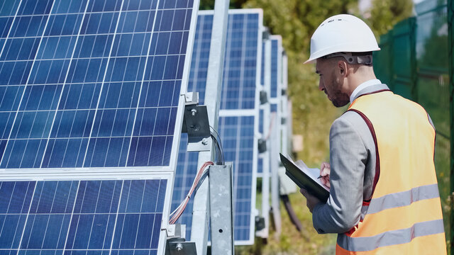 businessman in hardhat looking at wires and holding folder near solar batteries outside
