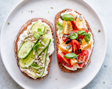 Two slices of toast with cream cheese, cucumber, tomato, pumpkin seeds, sesame seeds and basil