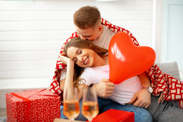 Young man hugs and kisses his beloved . They celebrate Valentine's Day at home in romantic setting with airy red hearts, gifts and wine.Love and care. Wonderful moments.