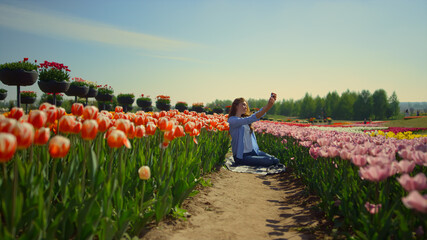 Young woman sitting in blooming flower garden with mobile phone in sunny day.