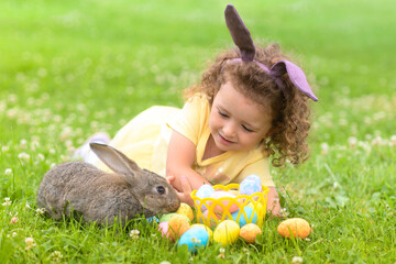 Happy Easter kid. little girl with very peri ears, small rabbit, grey bunny hunting for colorful eggs on green grass. spring holiday concept. copy space, text