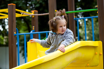 Fototapeta na wymiar Cute child girl with ponytails plays on the playground - runs, climbs, rides on a swing and a slide. Outdoor active leisure