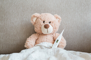 Brown teddy bear sitting on sofa under blanket and holding white digital thermometer. Temperature...