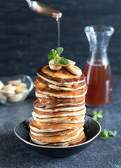 Maple syrup pours from a spoon onto a stack of pancakes with bananas and mint
