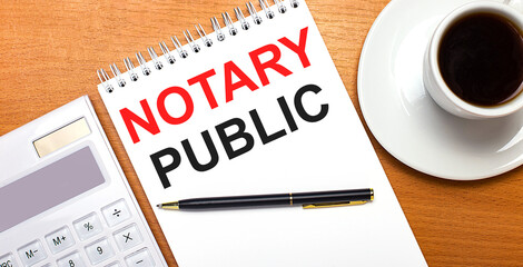 On a wooden table there is a white calculator, a white cup of coffee, a pen and a white notebook with the text NOTARY PUBLIC. Business concept