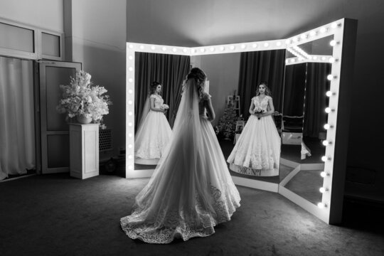 The bride in a white dress in front of the mirrors in the living room. Preparing for the upcoming wedding. Black and white photo.