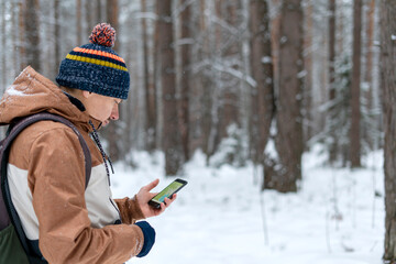 Young man in knitted hat with backpack looking at map on smartphone while hiking in winter forest...