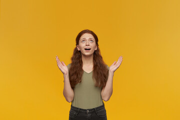 Indoor portrait of young ginger female keeps her finger crossed, looks upwards while praying for...