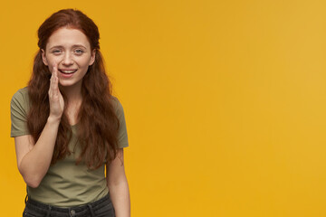 Indoor portrait of young ginger female whisper to her friend with happy facial expression,. isolated over yellow background