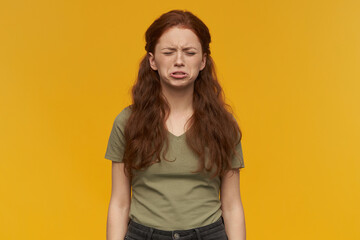 Fototapeta premium Indoor portrait of young ginger female keeps her eyes closed while crying. isolated over yellow background