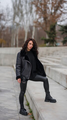 Girl on the steps. Stylish appearance. Total black. Girl with curly hair. Black jacket, black sweater, eco leather leggings.