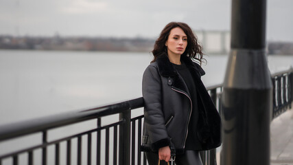 A girl in black clothes stands at the pier. Stylish look. Girl with curly hair. Against the background of the river.