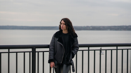 Obraz premium A girl in black clothes stands at the pier. Stylish look. Girl with curly hair. Against the background of the river.