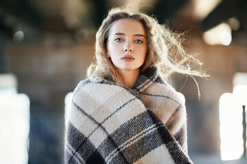 Close-up portrait of beautiful young woman wrapped in wool blanket looking to camera on windy sunny...