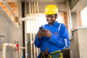 smilling young indian plumber or repairman busy by using mobile phone while working on industrial - concept of wasting of time by using social media, technology and internet distraction at workplace.