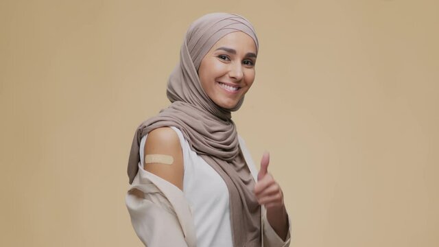 Young positive muslim lady wearing hijab showing shoulder with medical plaster after vaccination, gesturing thumb up