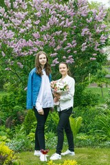 Two young beautiful girls, a portrait in the garden on a background of lilacs. Girls in a white blouse hold a beautiful bouquet of flowers in their hands.