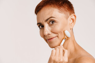Senior shirtless woman smiling while massaging face with jade roller