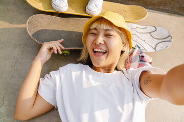 Asian girl laughing and taking selfie photo while lying on skateboard - obrazy, fototapety, plakaty