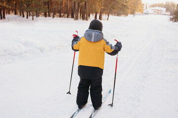 A little child in a yellow jacket cross-country skiing in the forest sliding down a snowy slope. Winter sport. A kid learns to ski. Active lifestyle, leisure. Sunny day