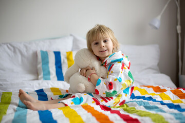 Happy toddler child, blond boy with colorful bathrobe, sitting in bed with stuffed toy after bath,...