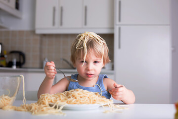 Little blond boy, toddler child, eating spaghetti for lunch and making a mess at home in kitchen