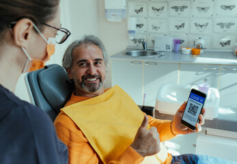 Smiling Male Patient Showing Thumbs Up and Vaccine Passport on his Mobile Phone App at Dentist's Office