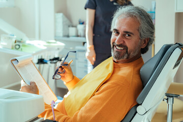 Portrait of Smiling Male Patient Filling Documents on a Clipboard while Sitting on a Dentist's Chair. 
Happy man looking at camera while holding anamnesis form fill and eyeglasses in dentist's office. - obrazy, fototapety, plakaty