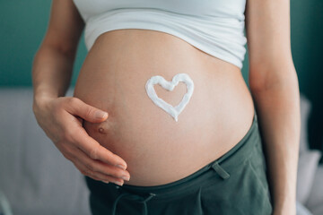 Close up of regnant woman dressed in light top and dark trousers hugging her belly with heart shaped cream on it symbolising love. Health and appearance care pregnancy, skin moistening and nourishment