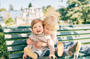 Outdoor portrait of two adorable siblings resting on the bench in sunny park, preschooler brother...