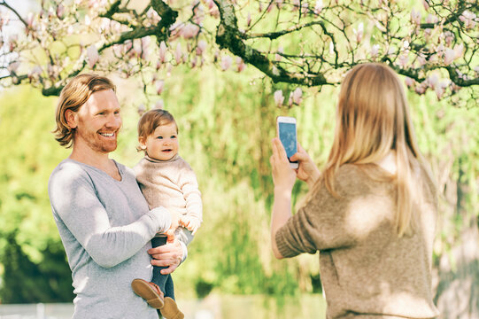 Young mother taking phone pictures of her husband and toddler daughter in spring park
