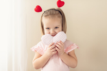 Obraz na płótnie Canvas Little girl holding heart. Small heart shape pillow holding by little Caucasian girl smiling with innocence, at home. Concept of child, love and happiness, kindness, gift, handmade Valentine, familly