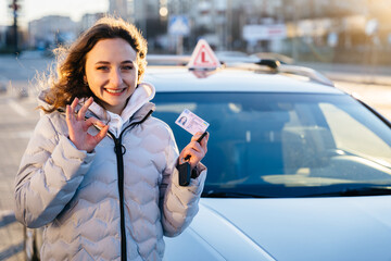 Young curly woman showing ok sign gesture and driving licence with examination car on background outdoor.