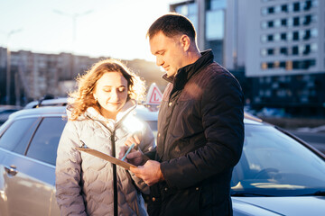 Happy young woman smiling and talking to male instructor with examination car on background. Driving instructor and woman student outdoor.